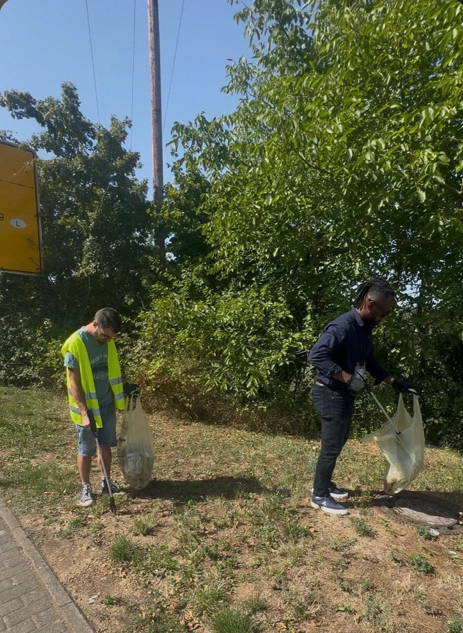 Deux personnes, dont une en gilet jaune, ramassent des déchets dans l'herbe au bord d'une route.