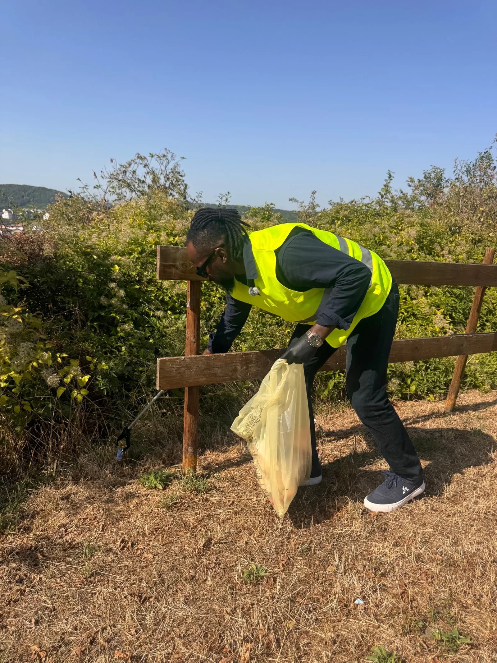Une personne en gilet jaune ramasse des déchets le long d'une barrière en bois.