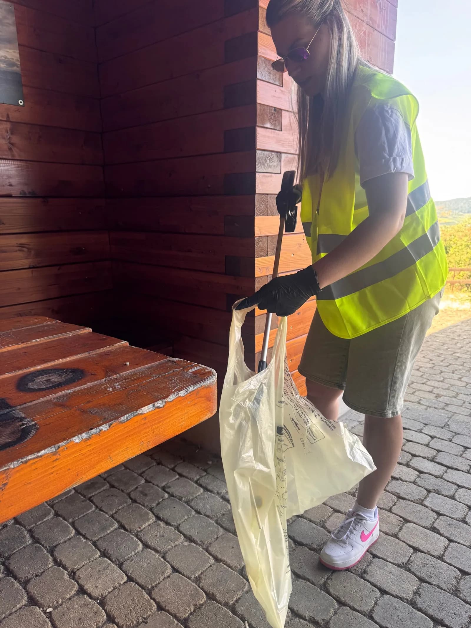Une femme en gilet jaune ramasse des déchets avec une pince près d'un abri en bois.