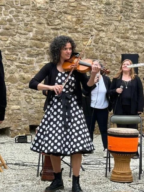 Une violoniste en robe à pois joue devant un mur de pierre, à côté de deux chanteuses et d'un djembé.