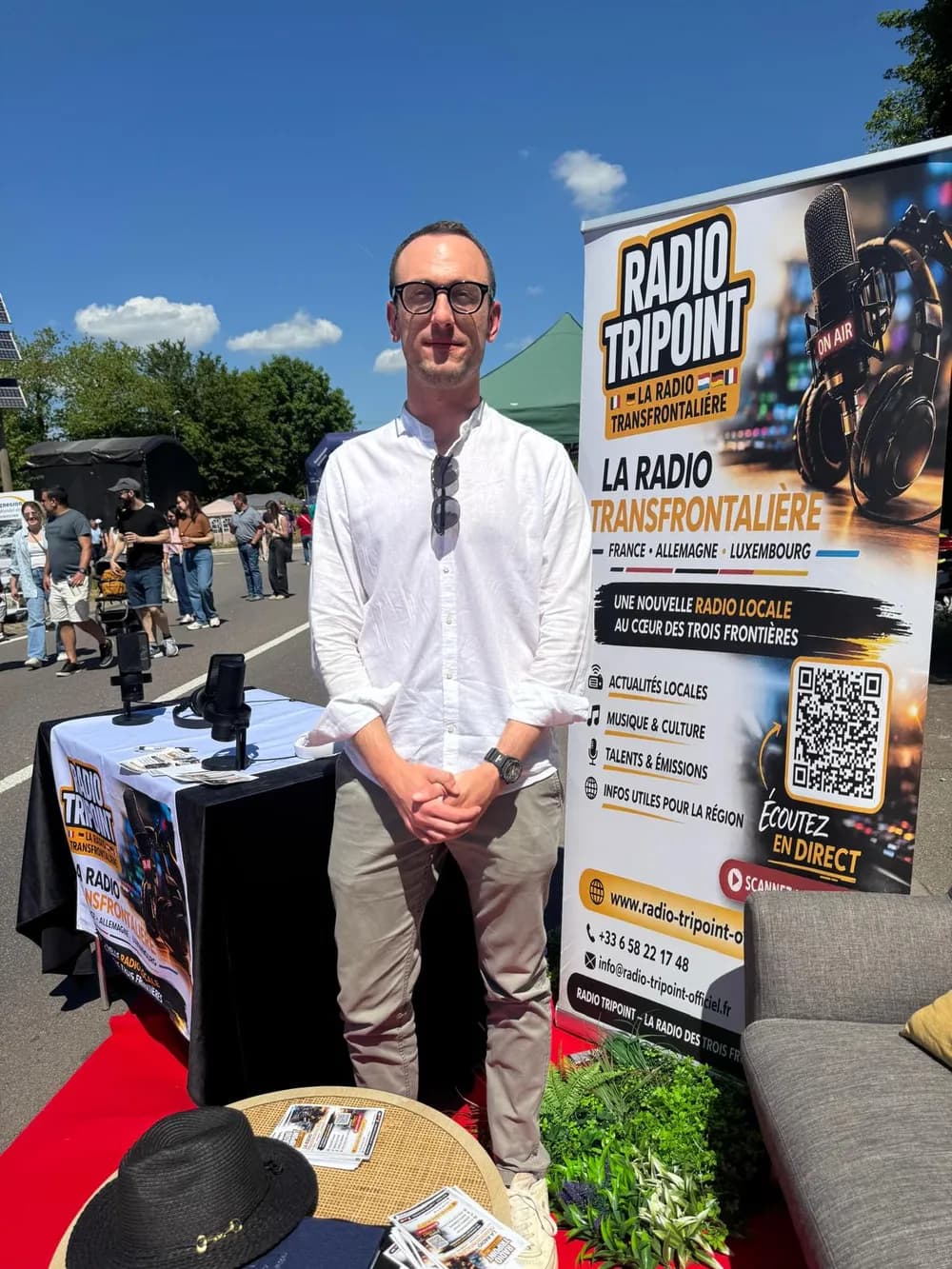 Lucas Grandjean, en chemise blanche, debout devant le stand de Radio Tripoint en plein air, à côté du kakémono de la radio.