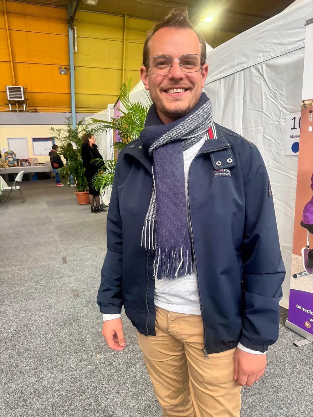 Rémy Dick, souriant, en blouson bleu marine et écharpe, dans une salle d'exposition entre des stands et des plantes vertes.